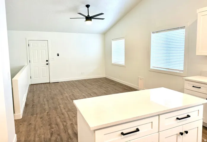 Vaulted ceiling with a ceiling fan, wood-finish flooring, multiple windows with blinds, and a kitchen island with white cabinetry and dark hardware