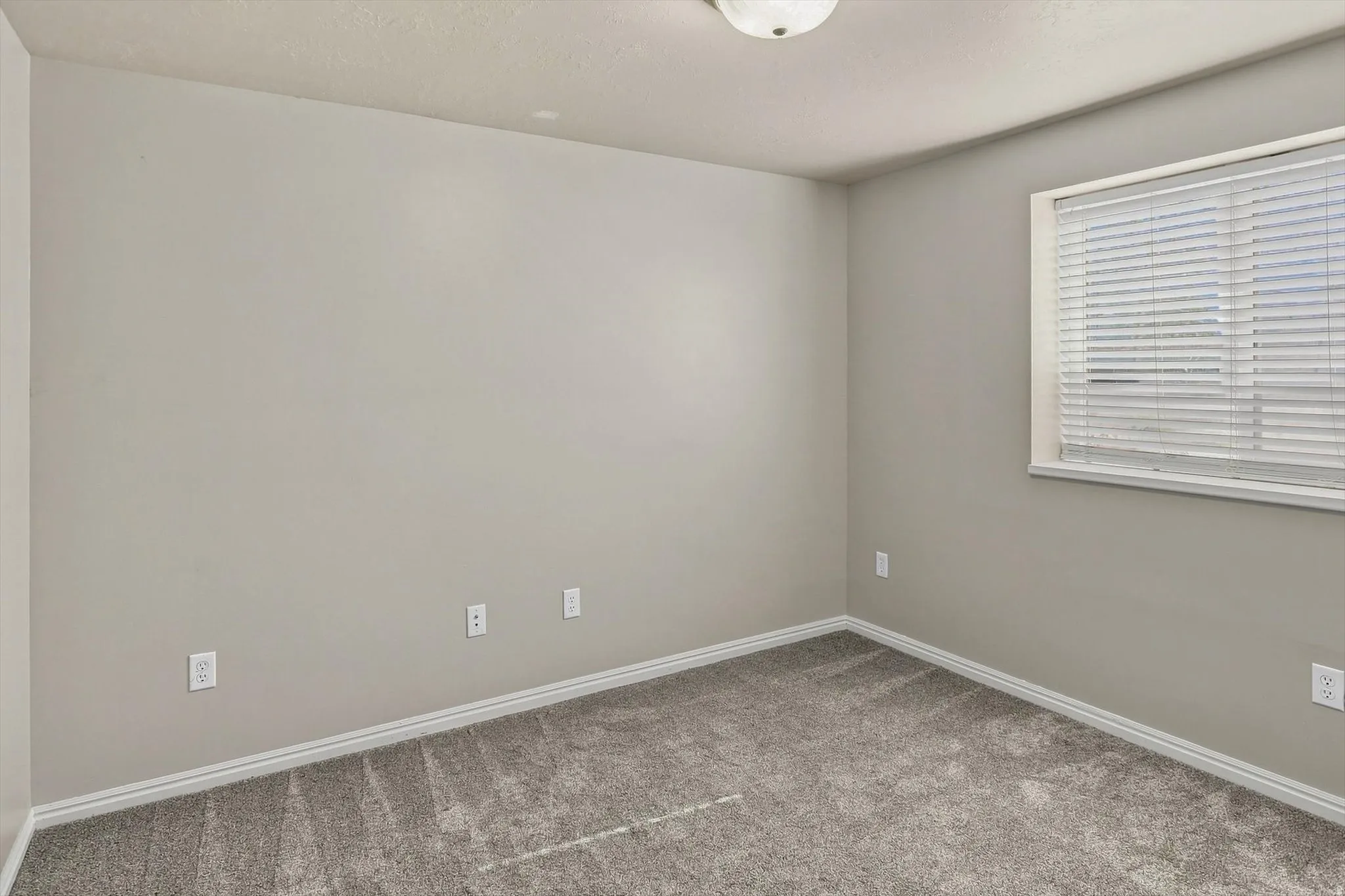 Bright, neutral bedroom featuring fresh carpet flooring, multiple electrical outlets for convenience, and abundant natural light from a large window with horizontal blinds