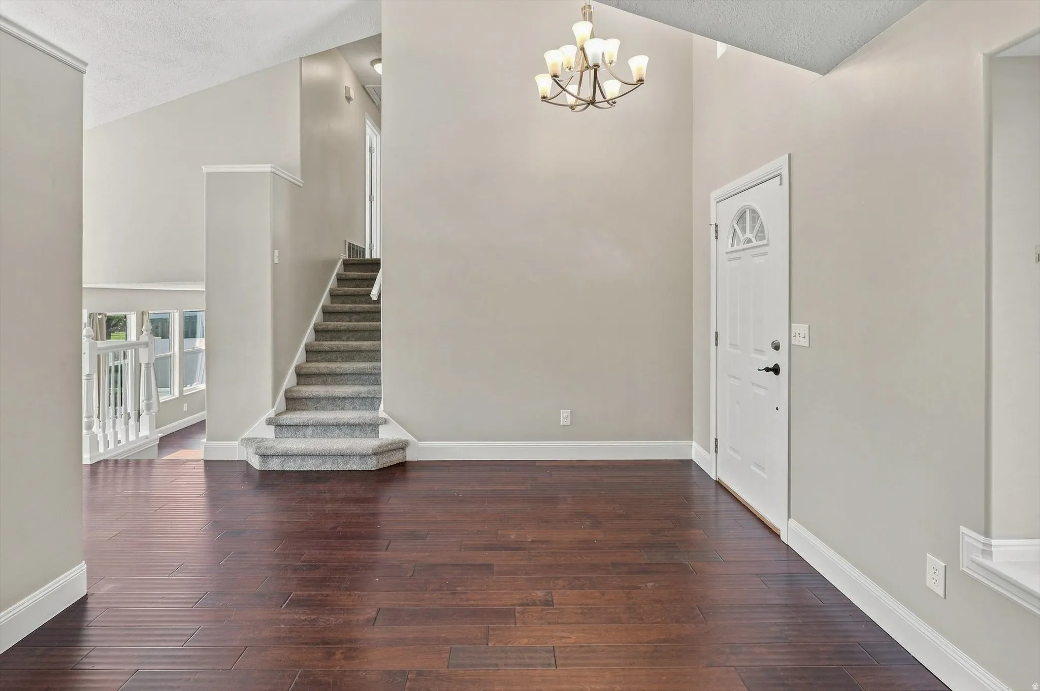 Vaulted ceiling with chandelier, dark wood-finish flooring, white trim, carpeted staircase, and an exterior door with an arched window