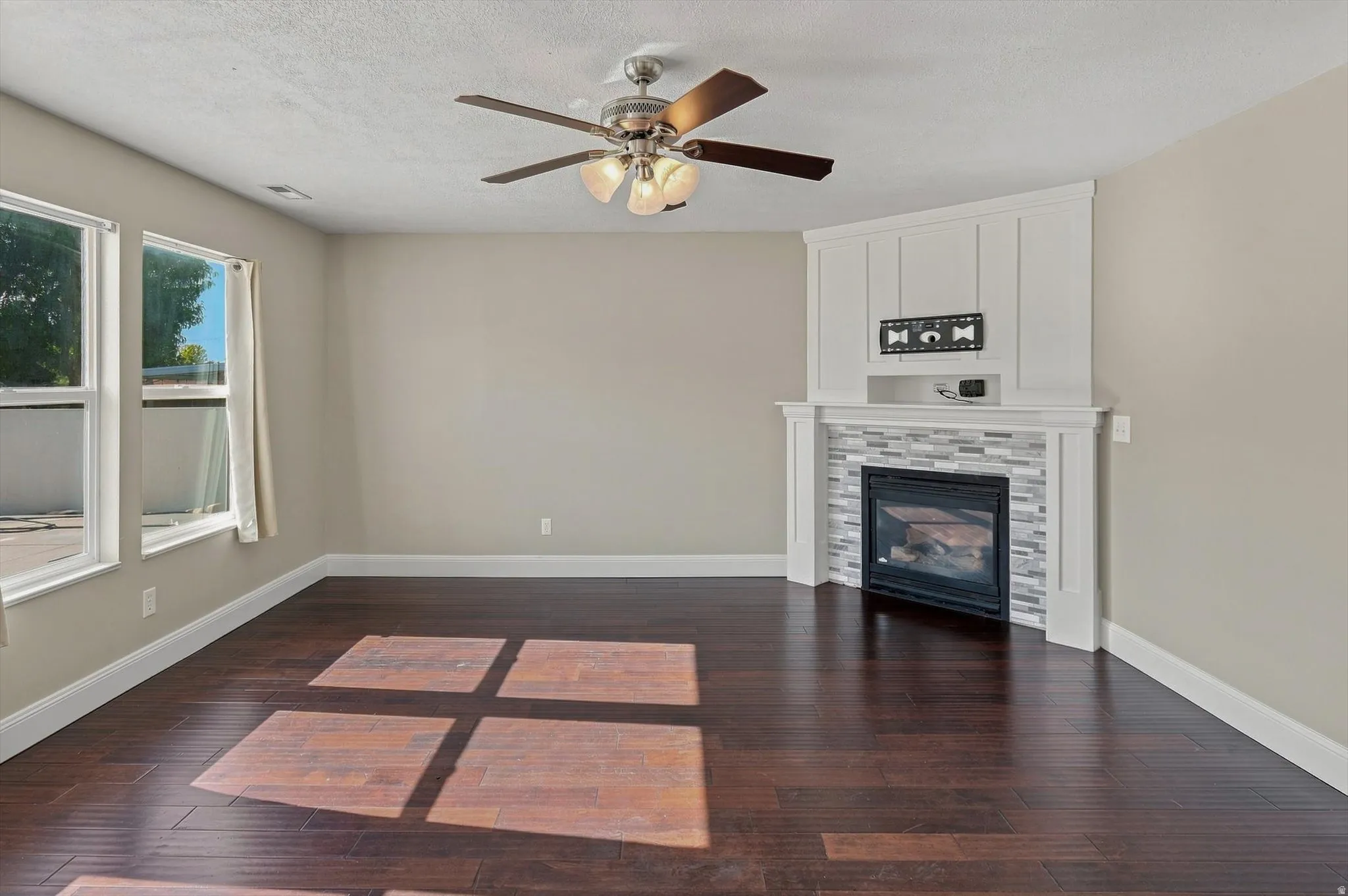 Hardwood flooring, corner fireplace with tile surround, built-in shelving, multiple windows, and a ceiling fan with integrated lighting