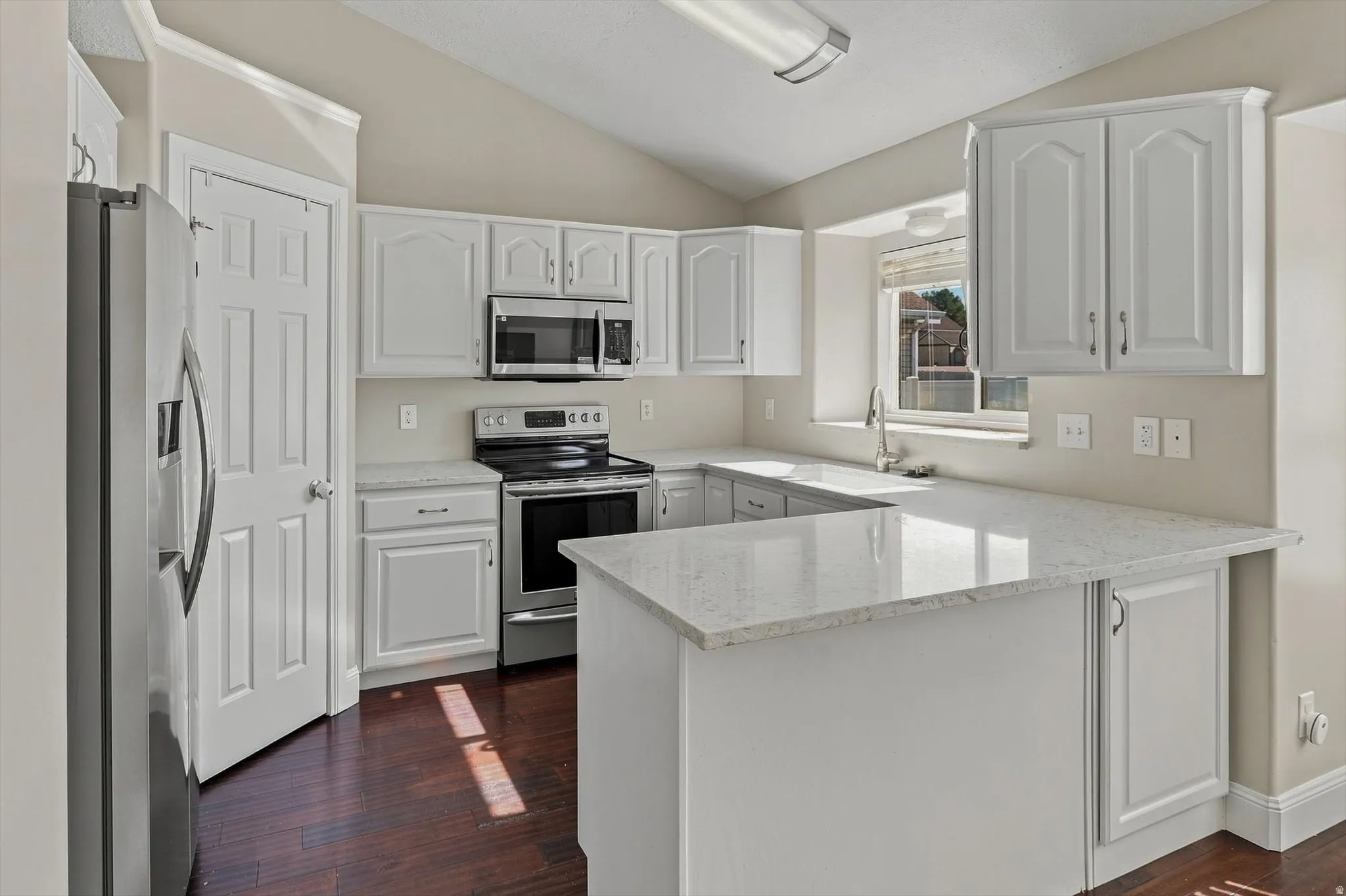 Bright, open kitchen featuring white cabinetry with raised-panel doors, stainless steel appliances including a microwave and electric range, quartz countertops, a center island with additional counter space, and wood-finish flooring throughout