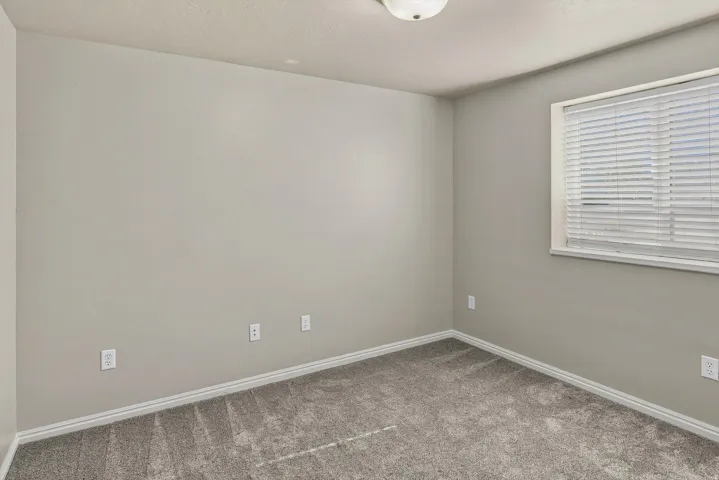 Bright, neutral bedroom featuring fresh carpet flooring, multiple electrical outlets for convenience, and abundant natural light from a large window with horizontal blinds
