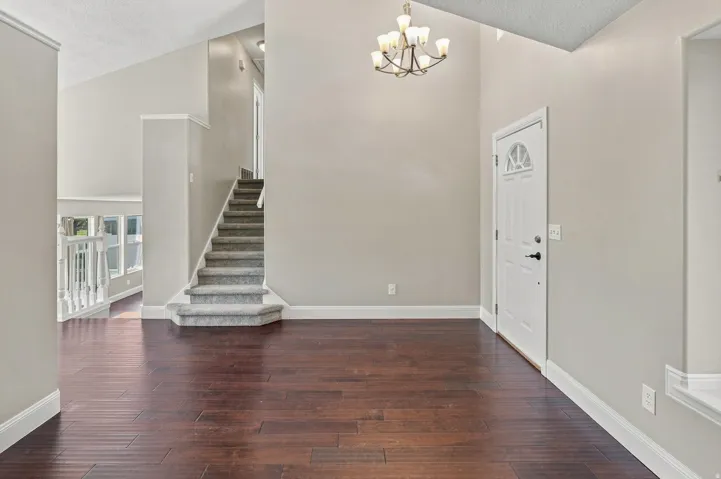 Vaulted ceiling with chandelier, dark wood-finish flooring, white trim, carpeted staircase, and an exterior door with an arched window