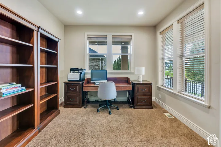 Dedicated home office featuring built-in wood cabinetry with open shelving, a wood desk with dual pedestals, recessed ceiling lighting, large multi-pane windows with plantation shutters, and neutral carpeting throughout