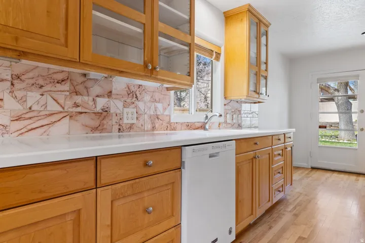 Wood-finish flooring, light wood cabinetry with glass-front uppers, white countertop, patterned tile backsplash, and a white dishwasher