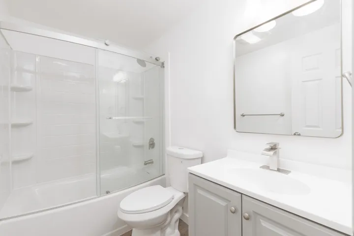 Bathroom featuring a tub and shower combination with a sliding glass enclosure, a vanity with a white countertop, and a wall-mounted mirror with overhead lighting