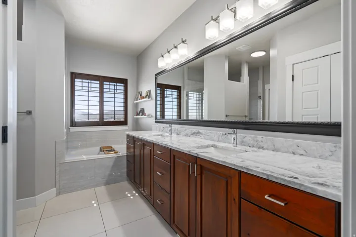 Dual vanity with stone countertops, wood cabinetry, built-in soaking tub, tile flooring, and wood plantation shutters