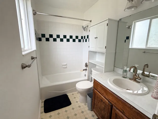 White tiled shower with a decorative checkered band, wood-finish vanity with a single basin sink, and a built-in white storage cabinet