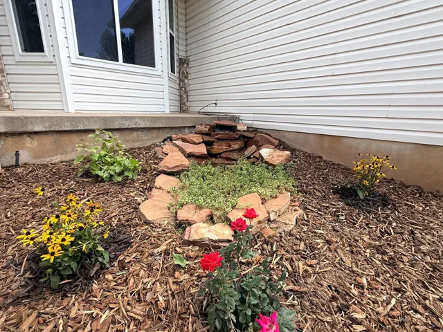 Landscaped garden bed with a rock water feature, varied plantings, and wood chip ground cover, set against white siding