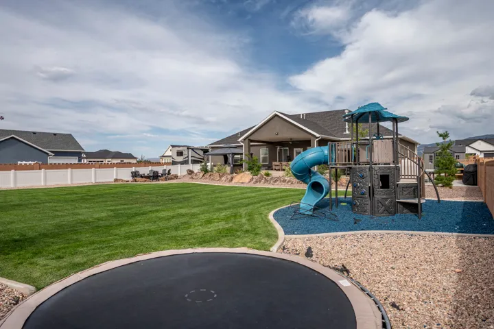 Expansive backyard featuring a manicured lawn, white vinyl fencing, an in-ground trampoline, and a dedicated play area with a structure and blue ground covering