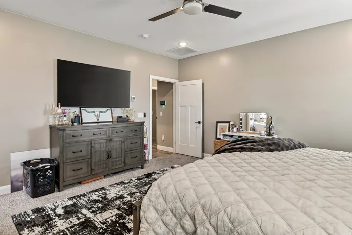 Spacious primary bedroom featuring a gray wood-finish dresser with ample storage, flat-screen television mounted above, ceiling fan with integrated lighting, and neutral wall finishes