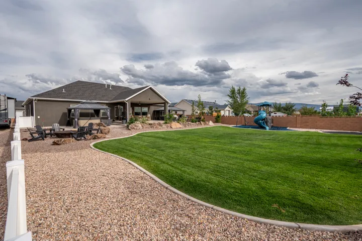 Expansive backyard featuring a lush lawn, gravel patio, covered outdoor living space, and a stone fire pit area
