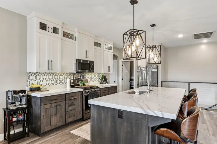 Contemporary kitchen and island featuring white shaker cabinetry with brushed metal hardware, dark wood-finish lower cabinets, white quartz countertops, stainless steel appliances, geometric tile backsplash, pendant lighting with geometric frames, and woo