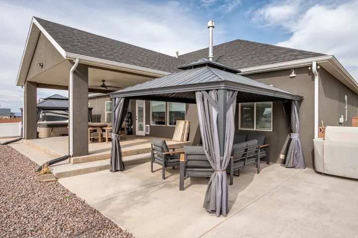 Expansive outdoor living space featuring a covered patio with integrated ceiling fan, adjacent pergola-style shade structure with curtain panels, and concrete hardscaping throughout