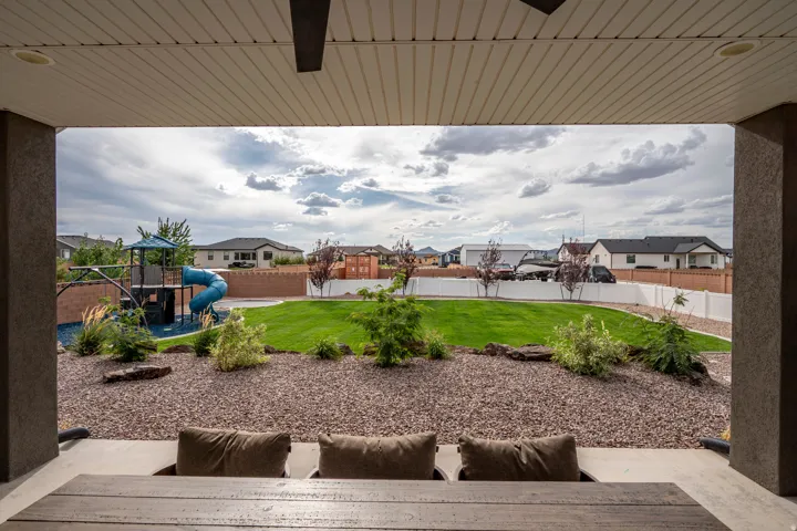 Covered patio with a ceiling fan, overlooking a spacious yard featuring a play structure, manicured lawn, and decorative rock landscaping