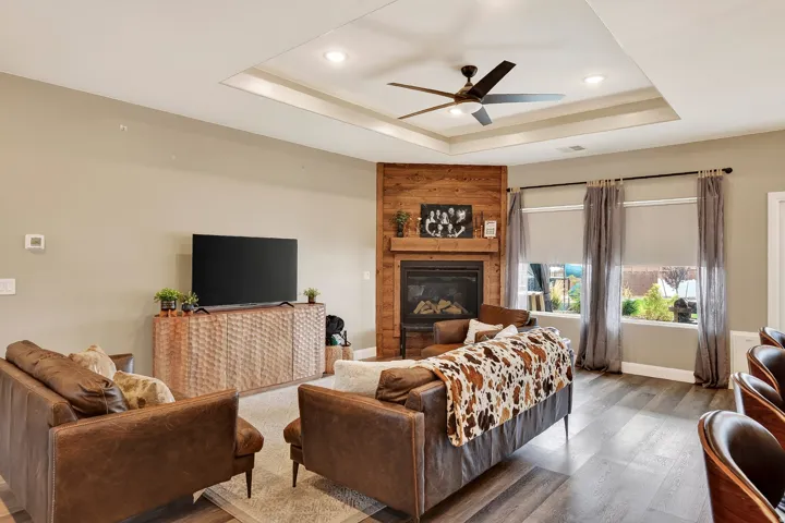 Living area featuring a gas fireplace with a wood-paneled surround, tray ceiling with recessed lighting, ceiling fan, wood-finish flooring, and multiple windows