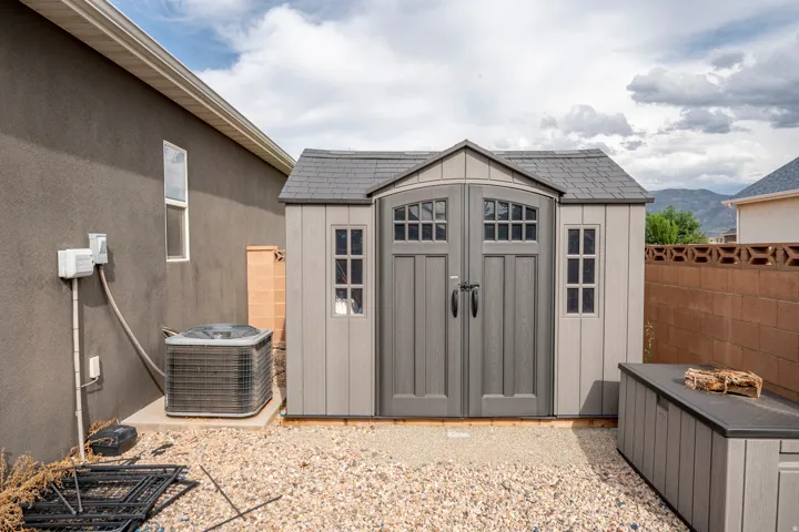 Gravel landscaping featuring a storage shed with dual doors and window accents, an air conditioning unit, and a concrete block privacy wall