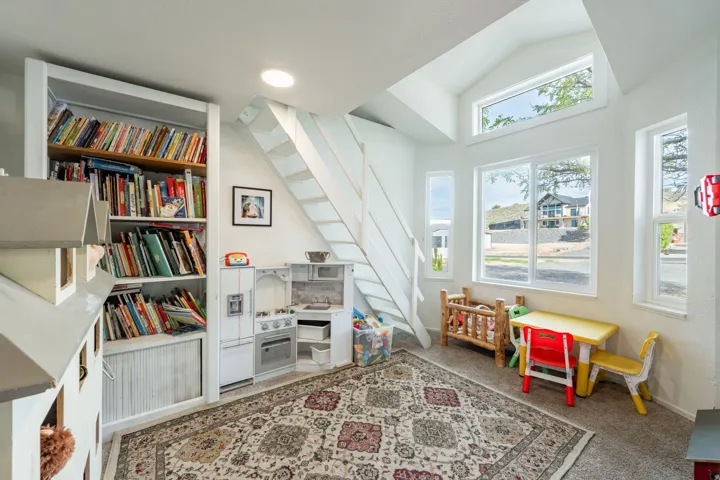 Bright, naturally lit room featuring built-in shelving with storage cabinetry, a skylight and multiple windows with natural light exposure, floating staircase with white railings, wood-finish flooring, patterned area rug, and a compact kitchenette with ap