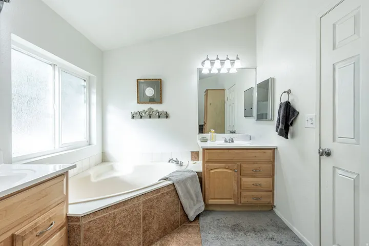 Soaking tub with tiled surround, dual vanity sinks with light wood cabinetry, and a frosted window