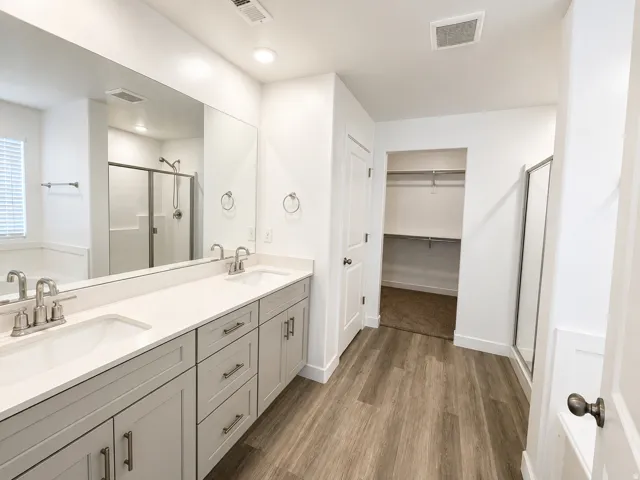 Dual vanity with light gray cabinetry and integrated sinks, wood-finish flooring, walk-in shower with sliding glass door, and a built-in closet with shelving
