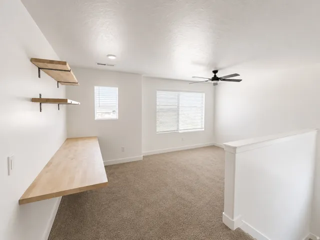 Built-in desk with wood-finish surface, floating wood-finish shelves, neutral carpet, and a ceiling fan