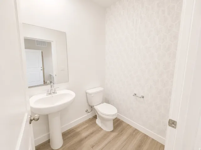 Pedestal sink with chrome faucet, toilet, wall-mounted mirror, geometric pattern accent wall, and wood-finish flooring