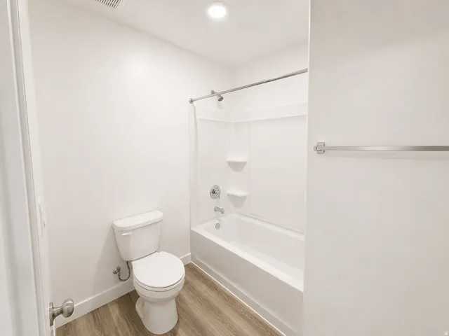 White acrylic bathtub and shower combination with chrome fixtures, wood-finish flooring, and a standard toilet