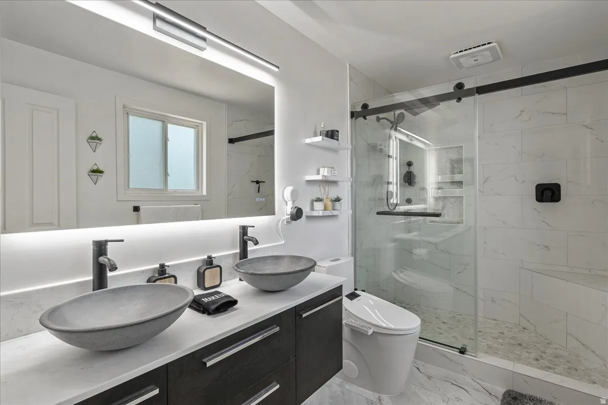 Dual vessel sinks, dark wood-finish vanity with drawer storage, back-lit mirror, glass-enclosed shower with tiled walls and built-in bench, and a wall-mounted toilet