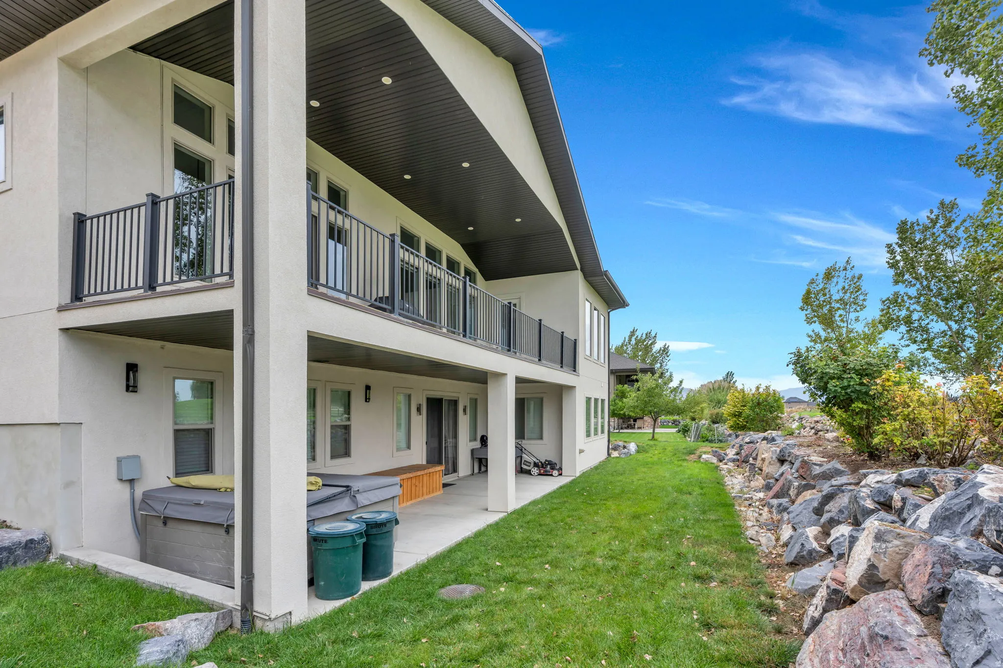 Multi-level exterior with a ground-level covered patio, upper-level balcony with black railings, and a landscaped rock retaining wall