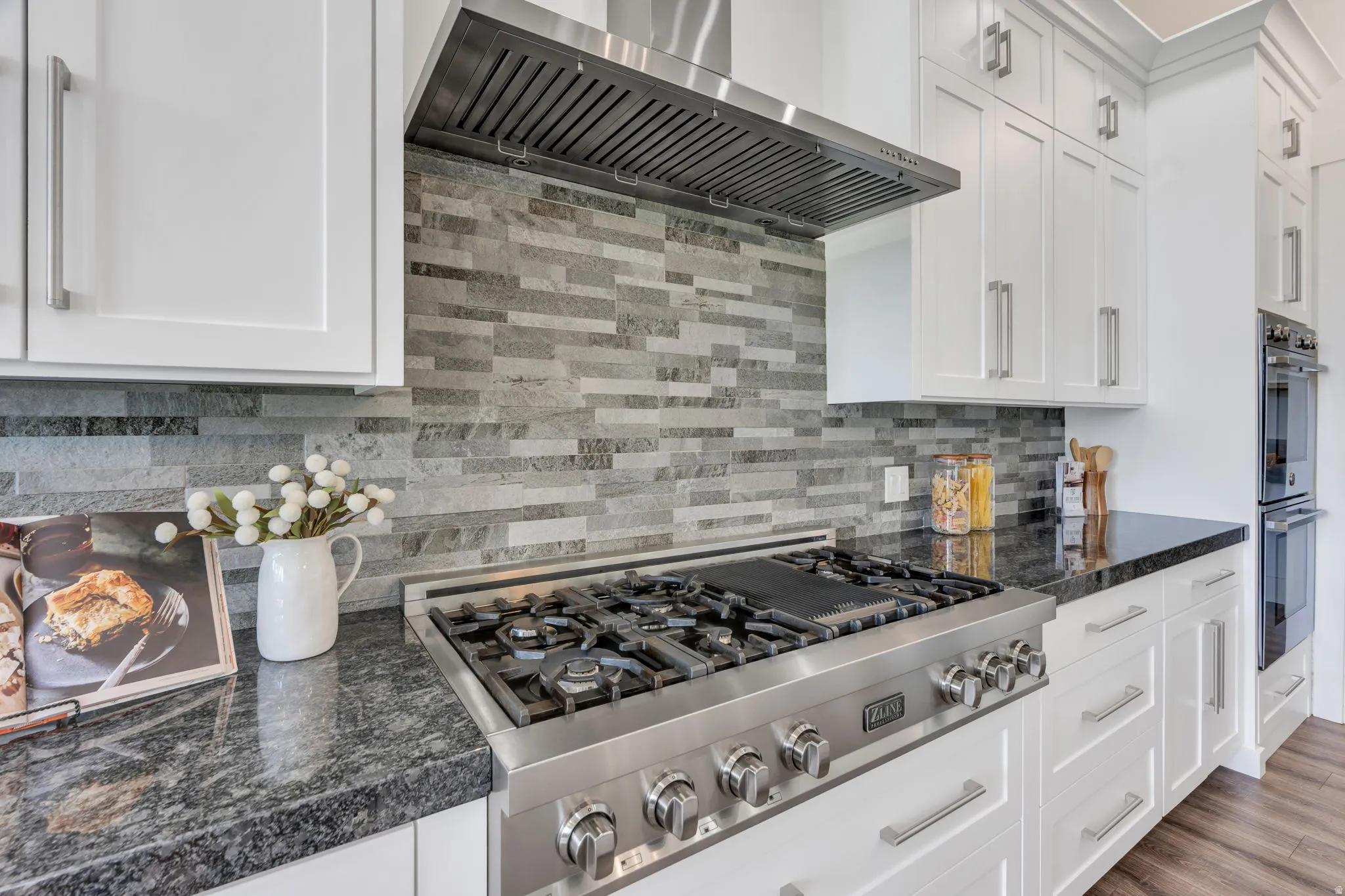 Stainless steel range with a ventilation hood, granite countertops, tile backsplash, white shaker-style cabinetry, and stainless steel double ovens