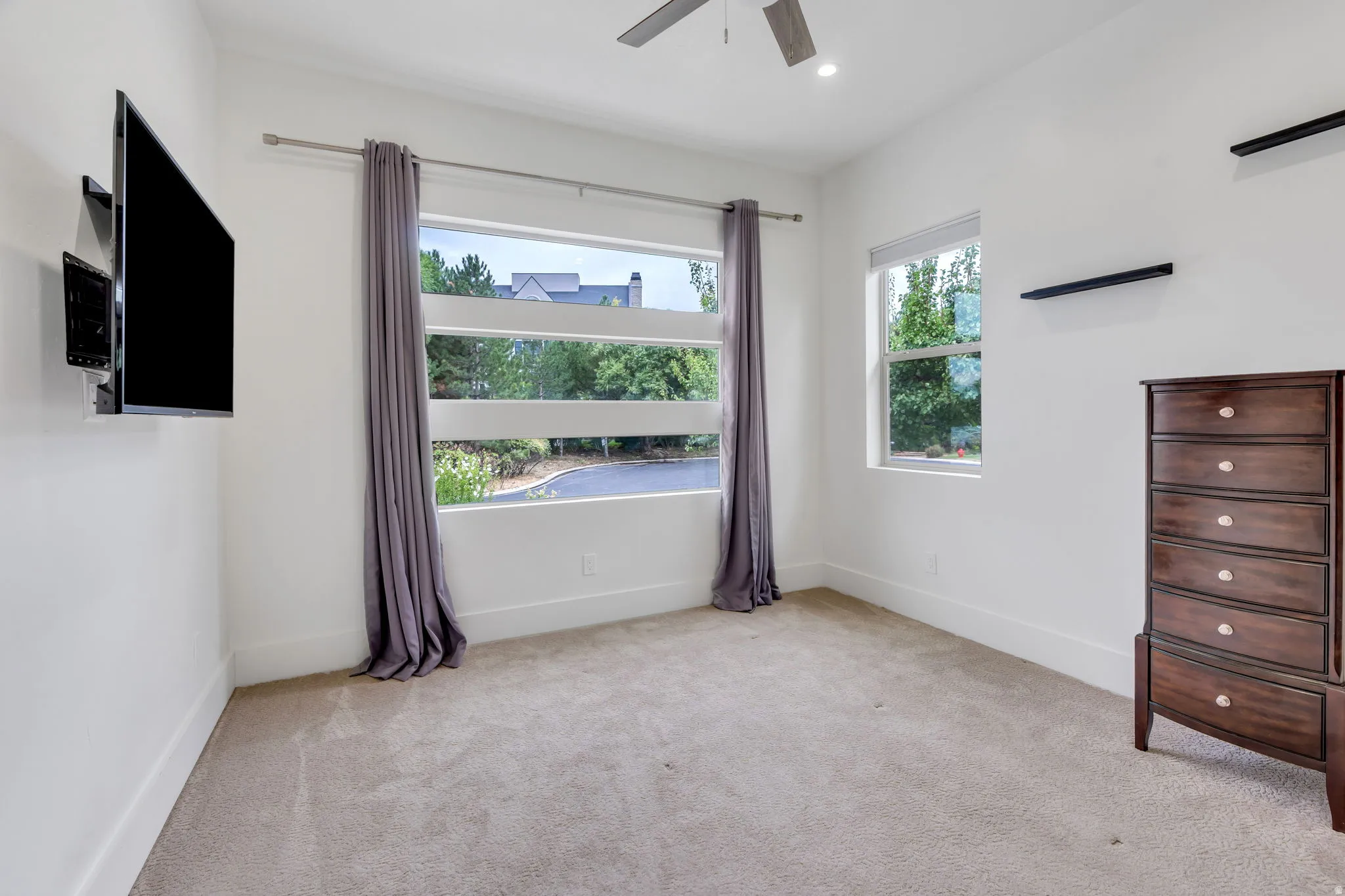 White walls, neutral carpeting, ceiling fan with integrated lighting, and two windows providing natural illumination