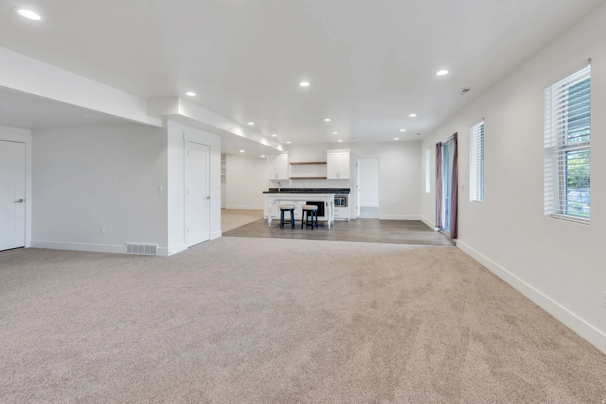 Recessed lighting throughout, two windows with blinds, a sliding glass door, a built-in kitchen area with white cabinetry, and wood-finish flooring