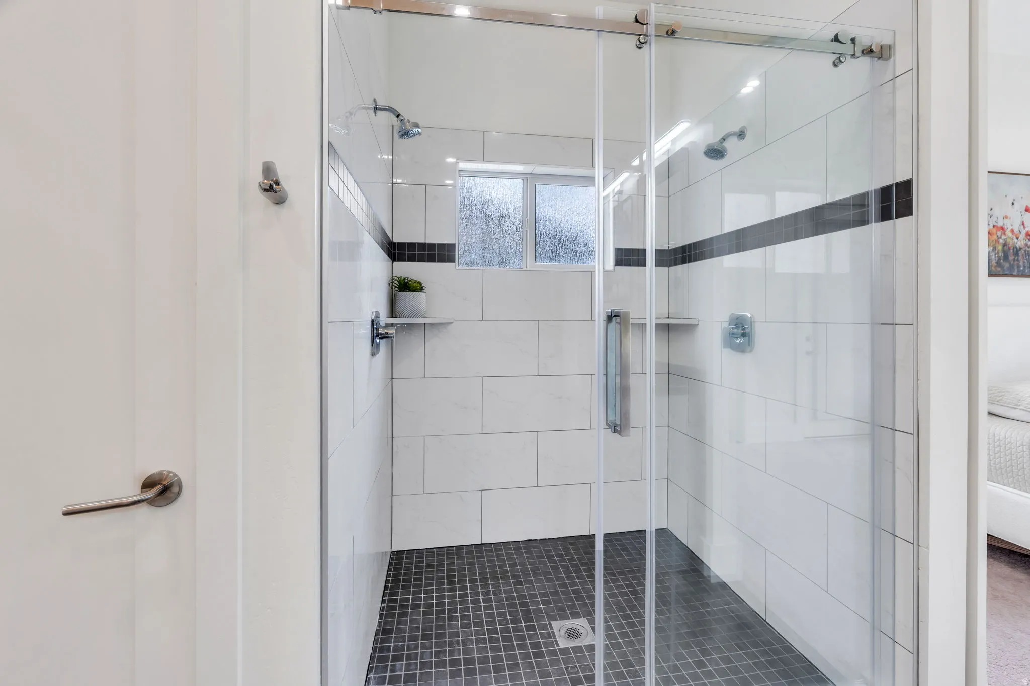 Frameless glass shower enclosure with sliding door, white subway tile walls, dark mosaic tile flooring, built-in shelving, and dual shower heads