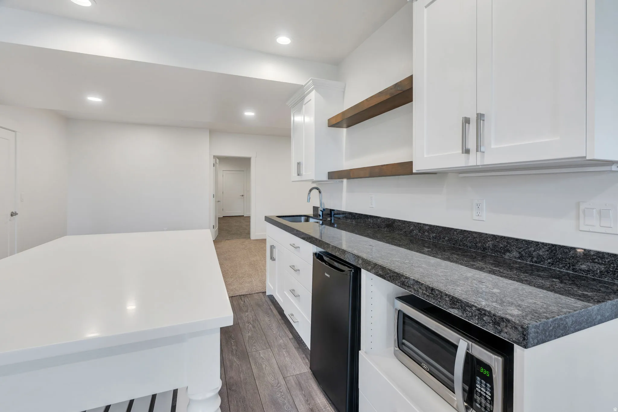 White cabinetry, dark stone countertops, stainless steel sink, wood-finish open shelving, and recessed lighting