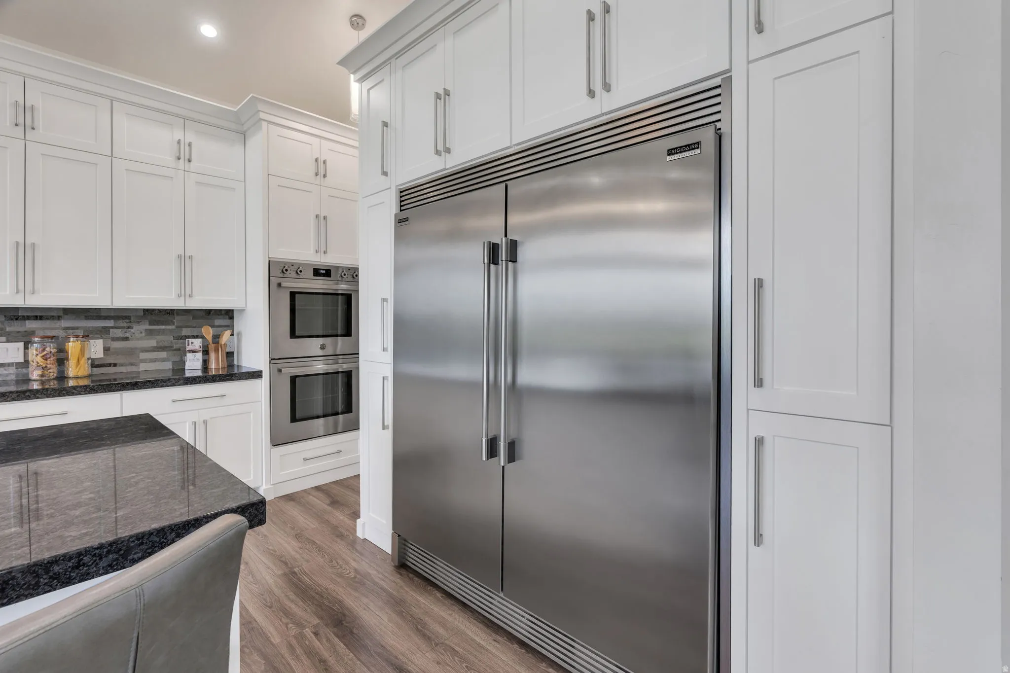 Built-in stainless steel refrigerator, double wall ovens, white shaker cabinetry, dark stone countertops, and wood-finish flooring