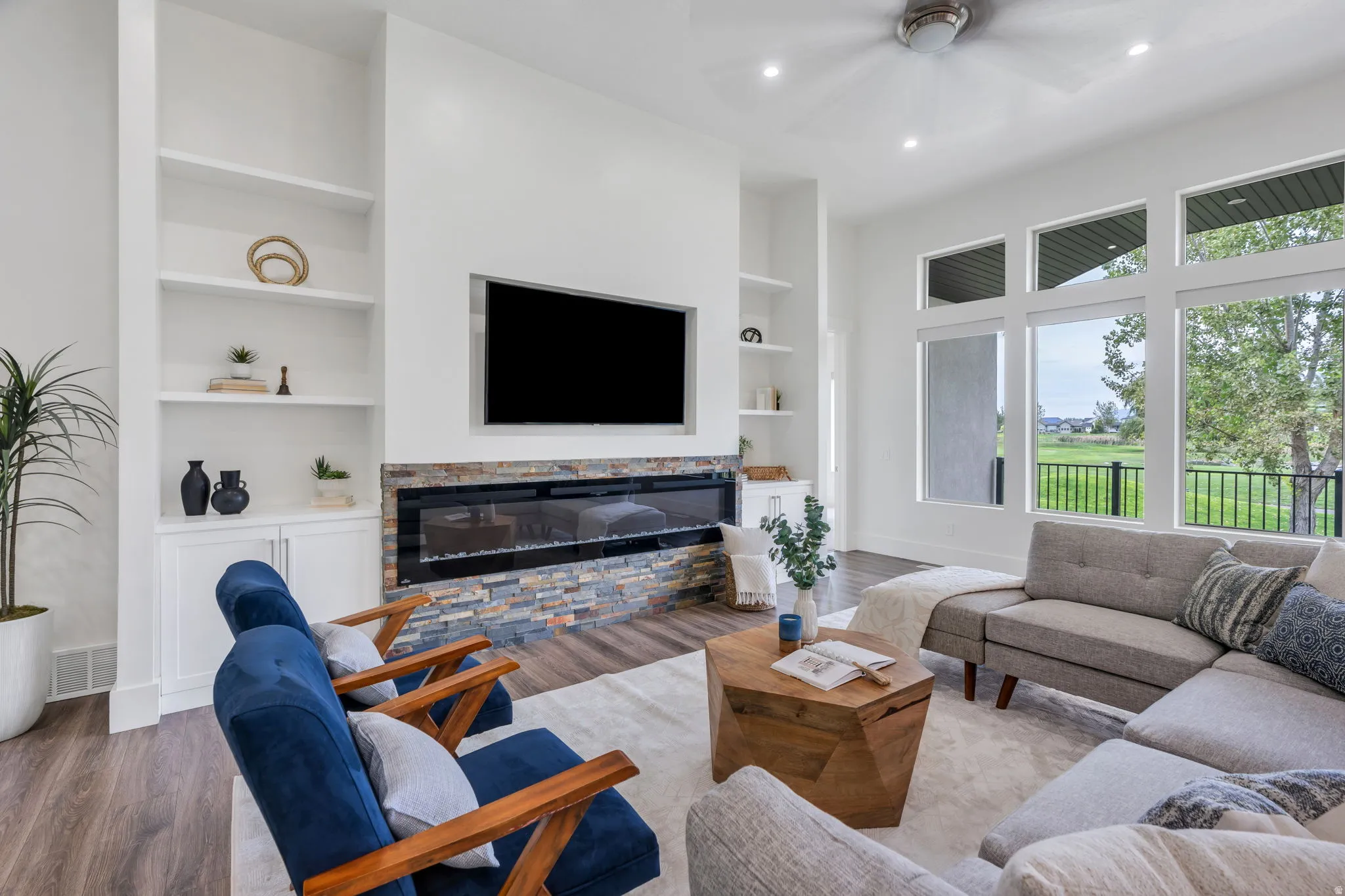 Open-concept living space featuring wood-finish flooring, a linear fireplace with stone surround, built-in shelving, and large windows with black exterior railings