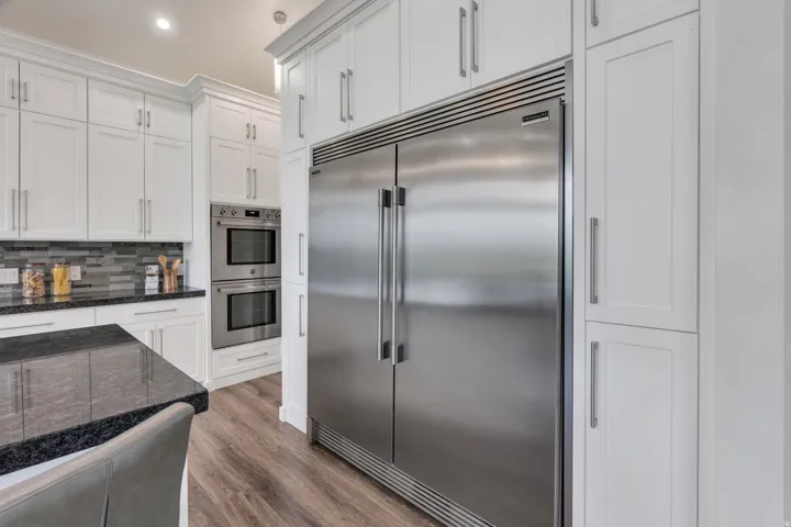 Built-in stainless steel refrigerator, double wall ovens, white shaker cabinetry, dark stone countertops, and wood-finish flooring
