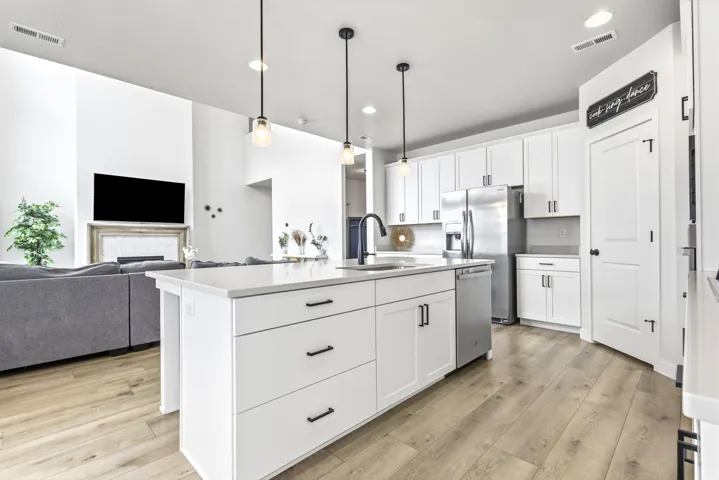 Open concept kitchen featuring an island with a built-in sink and pendant lighting, white cabinetry with black hardware, stainless steel appliances, and wood-finish flooring