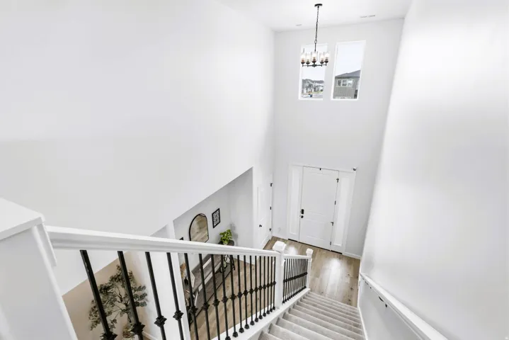 View from the tip of stairs to bottom with soaring ceilings with two upper windows, an elegant chandelier, a staircase with black wrought iron balusters, and a white entry door with sidelights.