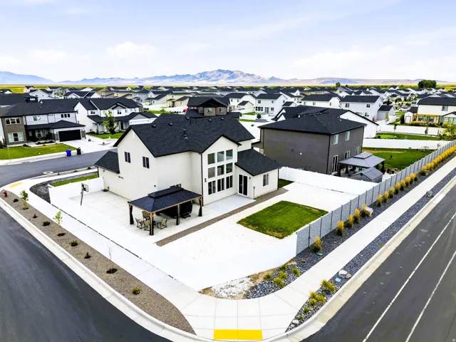 Modern exterior with white siding and dark roof, multiple large windows, corner lot positioning, concrete patio space, and surrounding neighborhood with mountain backdrop