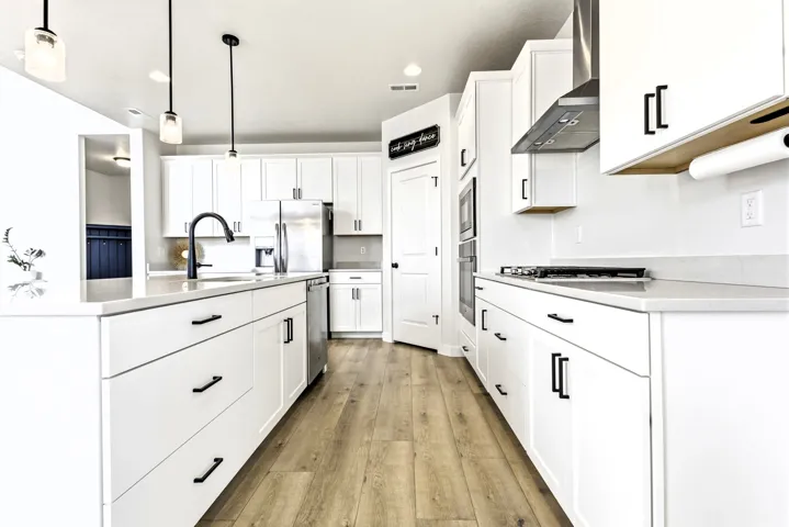 White cabinetry with matte black hardware, stainless steel appliances, light-toned wood-finish flooring, island with integrated sink, and pendant lighting