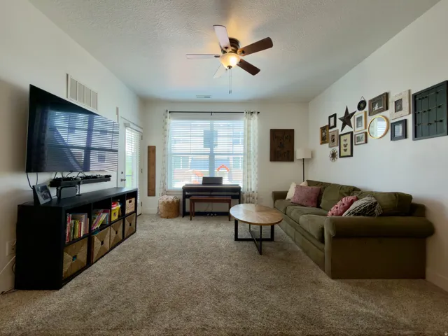 Spacious family room featuring carpeted flooring, a ceiling fan, a large window with blinds & a door to the covered deck