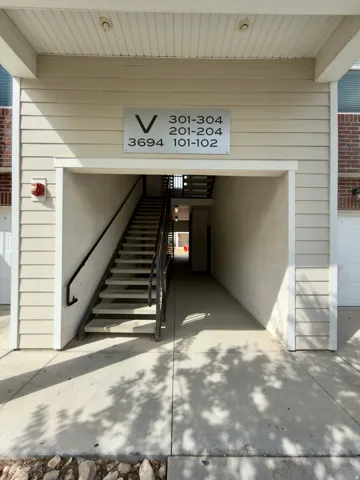 Exterior entry to building V with concrete walkway, a multi-level staircase with dark handrails, and a beige siding facade