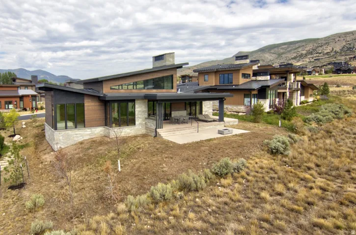 Modern architectural design featuring varied rooflines, extensive window placements, stone accent walls, wood-finish siding, and a spacious concrete patio