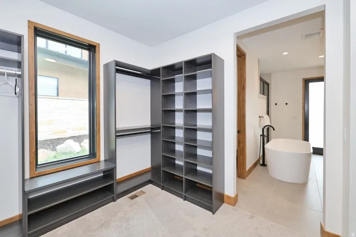 Built-in closet shelving and hanging rods, large vertical window with wood trim, and a standalone soaking tub with a floor-mounted faucet