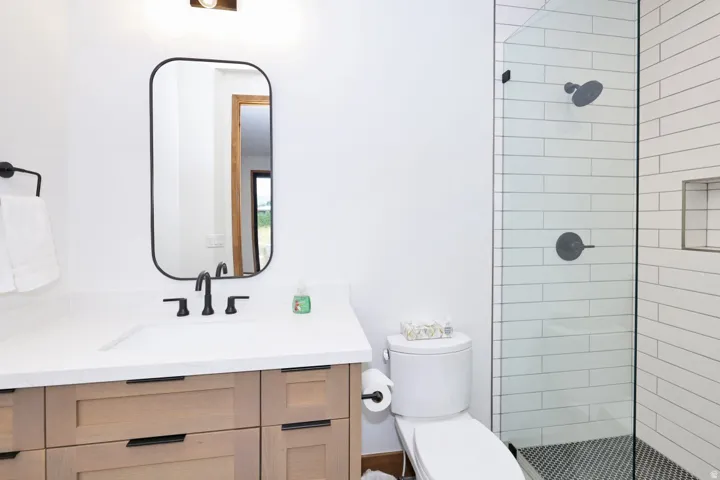 Wood-finish vanity with an integrated sink and matte black hardware, framed mirror, glass-enclosed shower with subway tile surround, and mosaic tile shower floor