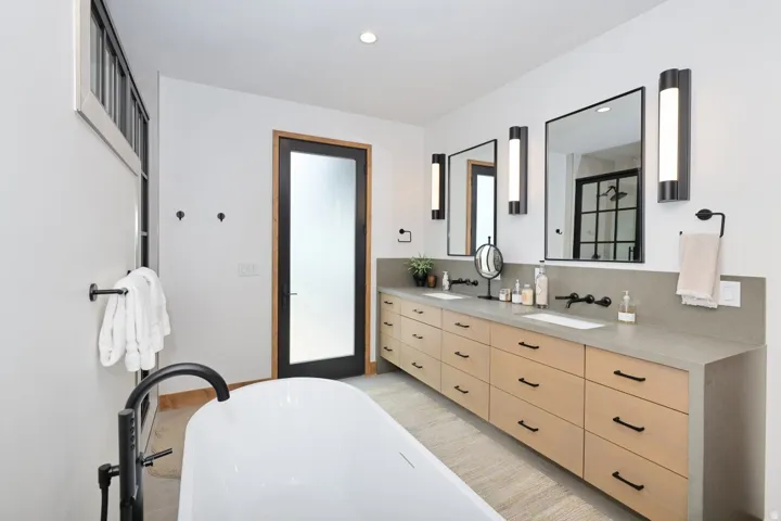 Spa-inspired primary bathroom featuring a freestanding soaking tub, dual vanity with wood-finish cabinetry and integrated storage, quartz countertops, vessel sinks, and matte black fixtures
