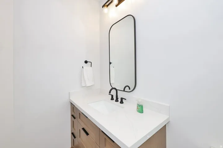Light wood-finish vanity with black hardware, white rectangular undermount sink, white countertop, and black-framed mirror