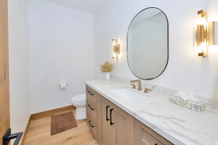 Light wood-finish flooring, a light wood-finish vanity with black hardware, a white countertop with gray veining, an oval mirror, and gold-tone wall sconces