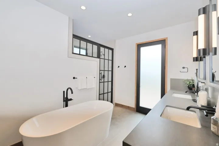 Spa-inspired primary bathroom featuring a freestanding soaking tub, dual vanity with quartz countertops and matte black fixtures, frameless glass shower enclosure with black metal detailing, recessed ceiling lighting, and direct access to an outdoor space